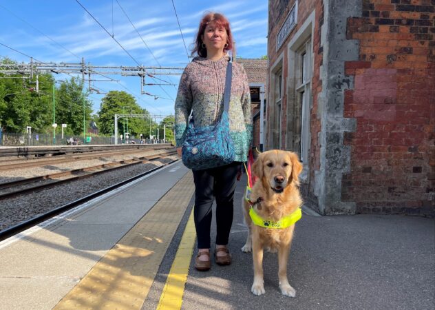 Photo shows Sarah Leadbetter, NFBUK’s National Campaigns Officer standing on a train platform with Nellie her Guide Dog in Harness. There is tactile paving and yellow line on the platform.